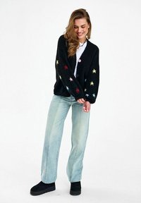 Žena oblečená v čiernom cardigane s farebnými vzormi hviezd, svetlomodrých širokých džínsach, čiernych topánkach, usmievajúca sa a pozerajúca sa dole.