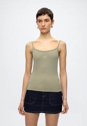 Γυναίκα με κοντά σκούρα μαλλιά που φοράει ασημένια σκουλαρίκια κρίκους, ελιά πράσινο καμησόλ με μικρό λογότυπο και σκούρη τζιν μίνι φούστα.