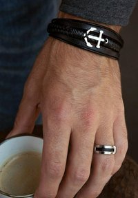 Zwart leren gevlochten armband met zilveren anker-achtige decoratie, gedragen om een hand, vergezeld van een zilveren ring en een kopje koffie.