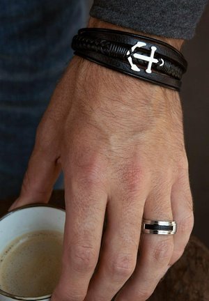 Zwart leren gevlochten armband met zilveren anker-achtige decoratie, gedragen om een hand, vergezeld van een zilveren ring en een kopje koffie.