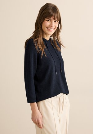 Marineblauer Hoodie mit weicher Textur, langen Ärmeln, Kordelzug am Hals und lockerer Passform, kombiniert mit hellen Hosen.