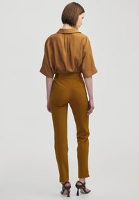 Braunes kurzärmliges Hemd mit weicher Textur, kombiniert mit hochtaillierten senfgelbfarbenen Hosen und schwarzen High Heels. Reduziertes Design.