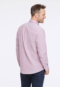 Camisa de manga larga rosa claro, confeccionada con tela texturizada. Presenta un cuello con botones y un dobladillo redondeado. Se lleva con jeans azul oscuro.