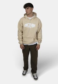 Felpa beige con scritta bianca "CRYSTAL PARIS," tasca a canguro, abbinata a pantaloni verde scuro e sneakers nere. Maglietta bianca visibile.