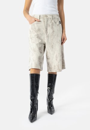 Persona con pantalones cortos hasta la rodilla de color gris claro con estampado y botas negras brillantes de tacón alto hasta la rodilla, de pie con una mano en el bolsillo.