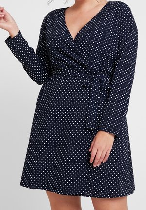 Vrouw draagt een marineblauwe wikkeljurk met lange mouwen en witte polkadots, gestrikt in de taille met een bijpassende stoffen riem.
