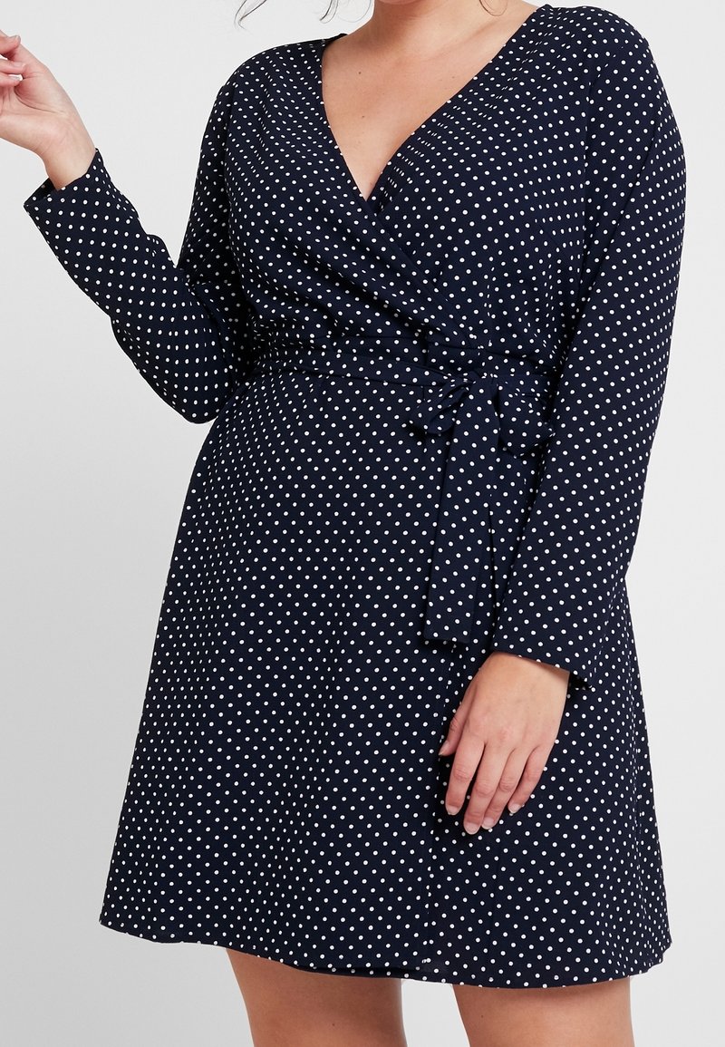 Femme portant une robe portefeuille bleu marine à manches longues avec des pois blancs, nouée à la taille avec une ceinture en tissu assorti.