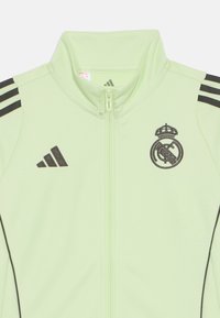 Lichtgroene zip-up tracksuitjack van zacht materiaal. Bevat zwarte adidas-strepen op de schouders en het Real Madrid-logo in zwarte borduurwerk.