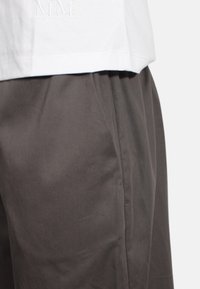 Chemise blanche à manches courtes avec des initiales brodées "MM" associée à un pantalon gris au tissu lisse et à la coupe cintrée.