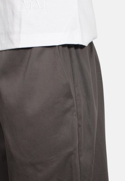 Bílá košile s krátkým rukávem, na které jsou vyšitá iniciály "MM", kombinovaná se šedými kalhotami se hladkým povrchem a úzkým střihem.