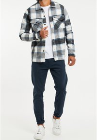 Camicia a scacchi di flanella bianca, blu e grigia; due tasche sul petto, design con bottoni, indossata sopra una t-shirt bianca e pantaloni blu navy, con sneaker bianche.