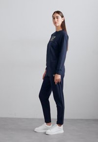 Tumši zils džemperis un bikses kopums no mīksta auduma. Džemperim ir logo iezīme; biksēm ir sašaurināts piegriezums un ērts elastīgs jostasvīrs.