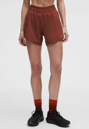 Persoon draagt roestkleurige atletiekshorts, oranje gradient sokken en zwarte hardloopschoenen, staand op een effen witte achtergrond.