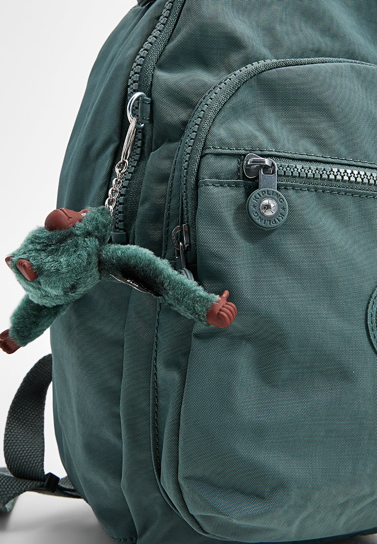 Mochila de tela verde con un bolsillo con cremallera en la parte delantera y un llavero de mono de peluche sujeto al tirador de la cremallera lateral.