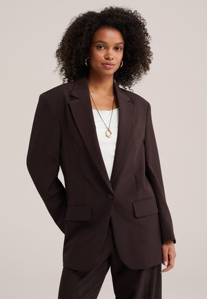 Donna con capelli ricci indossa giacca e pantaloni da completo marrone scuro sopra una maglia bianca, accessoriati con orecchini a cerchio dorati e una collana con ciondolo.