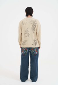Beige gerippter Pullover mit grafischen Drucken auf dem Rücken. Blaue weit geschnittene Jeans mit buntem Drachen- und Blumen-Stickerei auf der Rückseite.