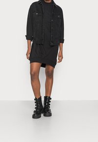 Černá denimová bunda navlečená přes černé šaty s kulatým výstřihem a volným střihem. Černé kozačky po kolena s několika sponami.