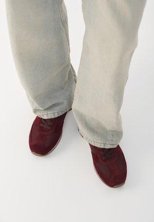 Sneakers in suede bordeaux con accenti in rete, con lacci frontali e suole in gomma, abbinati a jeans azzurri chiari, leggermente sfrangiati in fondo.