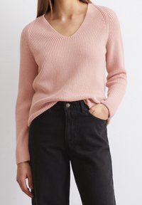 Un pull v-neck à côtes de couleur rose clair, à manches longues, porté avec un jean noir taille haute. Le pull présente une coupe décontractée et un tissu texturé.