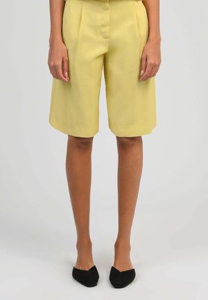 Personne portant un short jaune mi-cuisses et des chaussures plates noires à bout pointu, debout devant un fond clair uni.