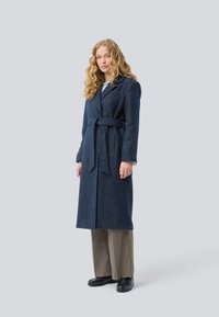 Frau mit langen, lockigen Haaren, die einen taillierten, marineblauen Wollmantel, graue weite Hose und schwarze Loafers trägt, steht vor einem schlichten, hellen Hintergrund.