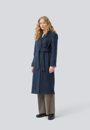 Mulher com cabelo longo e encaracolado, vestindo um casaco de lã azul-escuro com cinto, calças largas cinzentas e mocassins pretos, de pé contra um fundo claro e liso.