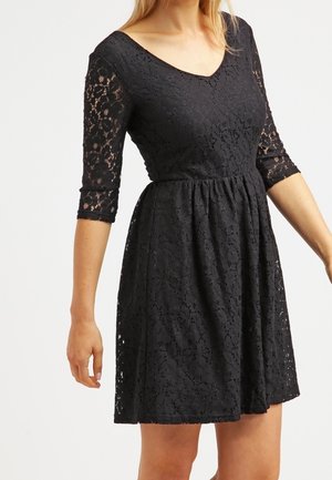 Frau trägt ein schwarzes Spitzenkleid mit Dreiviertelärmeln und geraffter Taille, steht vor einem schlichten hellen Hintergrund.