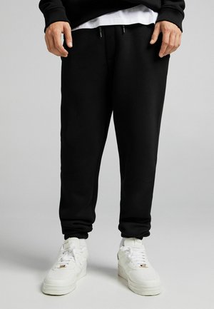 Homme portant un pantalon de jogging noir et des baskets blanches, debout devant un fond gris uni.