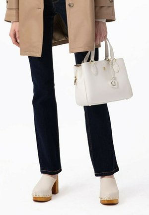 Wit leren handtas met dubbele handvaten, gouden accenten en een hangende charme. Draagt bij donkere jeans en beige klompen met een houten zool.