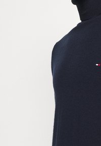 Dunkelblauer Rollkragenpullover aus weichem Strickstoff. Präsentiert ein kleines besticktes Logo in Rot, Weiß und Blau auf der Brust.