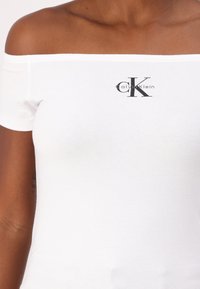 White off-the-shoulder fitted t-shirt made of cotton, featuring a black Calvin Klein logo at the center. Smooth texture and simple design.