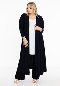 Langer, navyfarbener Cardigan aus weichem Stoff, mit lockerer Passform und offenem Vorderteil, getragen über einem weißen Oberteil und schwarzen weitem Hosen. Weiße Sandalen.