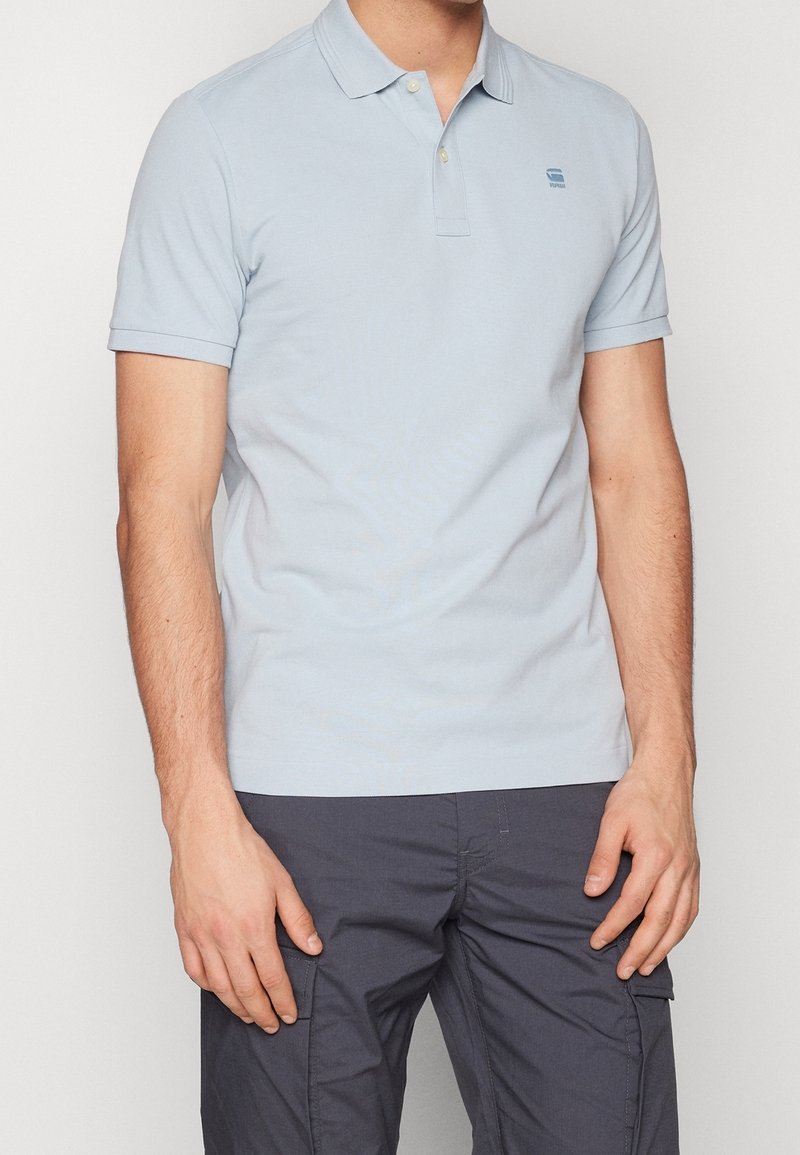 Homme portant un polo bleu clair à manches courtes et un pantalon cargo gris foncé, debout avec les bras détendus sur fond blanc.