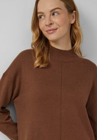 Brauner Strickpullover mit hohem Stehkragen, gerippter Struktur, überschnittenen Schultern und mittlerer Naht. Weicher Stoff mit lockerem Schnitt.