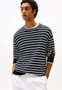Maglione a righe blu navy e bianche con scollo a girocollo e polsini arricciati, con un piccolo logo sul petto. Indossato con pantaloni bianchi.
