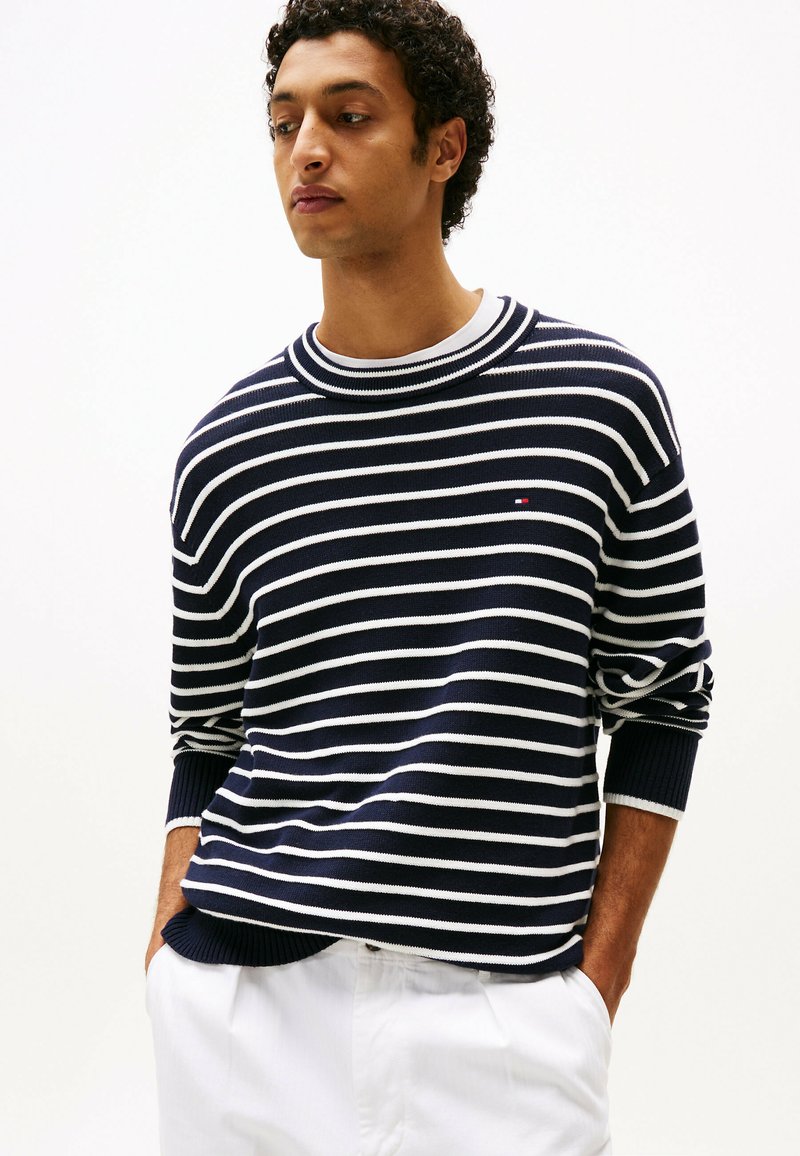Maglione a righe blu navy e bianche con scollo a girocollo e polsini arricciati, con un piccolo logo sul petto. Indossato con pantaloni bianchi.