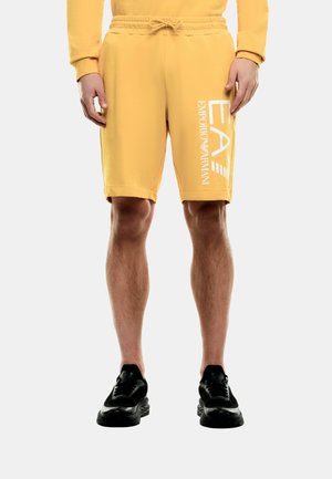 Hombre vistiendo pantalones cortos amarillos con cordón y el logo blanco "EA Emporio Armani" en el muslo derecho, y zapatos deportivos negros.