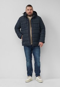 Mężczyzna ubrany w granatową pikowaną kurtkę, beżowy sweter, niebieskie dżinsy i białe sneakersy stoi na jednolitym jasnoszarym tle.