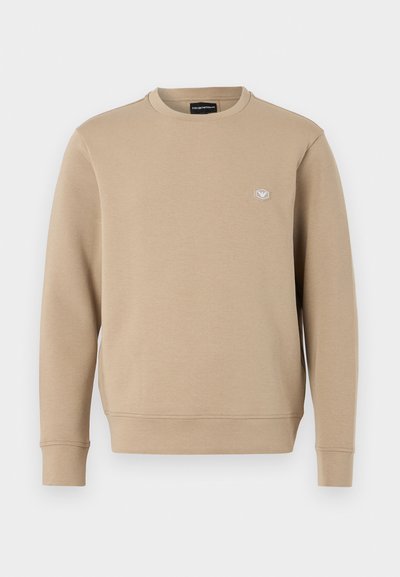 Beige crew neck sweatshirt tillverkad av mjukt tyg. Har en liten logopatch på bröstet. Långa ärmar med ribbade muddar och nederkant.