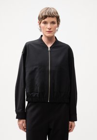 Veste bombardier noire en tissu lisse, dotée d'une fermeture éclair à l'avant, de deux poches avant et d'un ourlet froncé pour un ajustement décontracté.