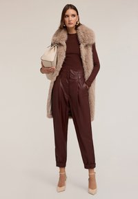 Lange, beige nepbont vest over een maroon ribgebreid shirt en maroon high-waisted leren broek. Lichtbeige tas en crèmekleurige hakschoenen.