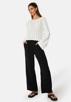 White knitted pullover with vertical stripes, wide sleeves; paired with black wide-leg pants; model wearing black flip-flops, hands in pockets.