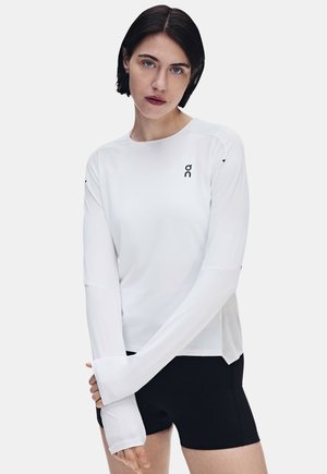 Frau mit kurzen dunklen Haaren, die ein weißes Langarmshirt und schwarze Shorts trägt, steht vor einem schlichten hellen Hintergrund.