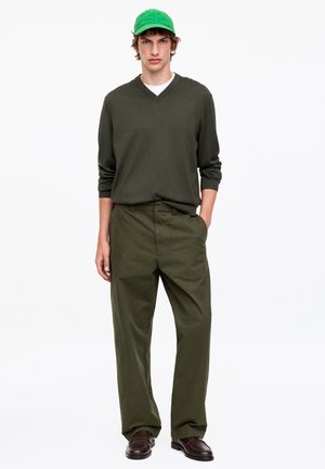 Homme debout face à l’avant, portant une casquette de baseball verte, un pull en V vert foncé, un pantalon vert olive et des chaussures marron sur fond blanc.