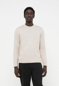 Suéter de punto beige claro con un cuello redondo y mangas largas, que presenta una textura suave y puños y dobladillo acanalados. Combinado con pantalones negros.