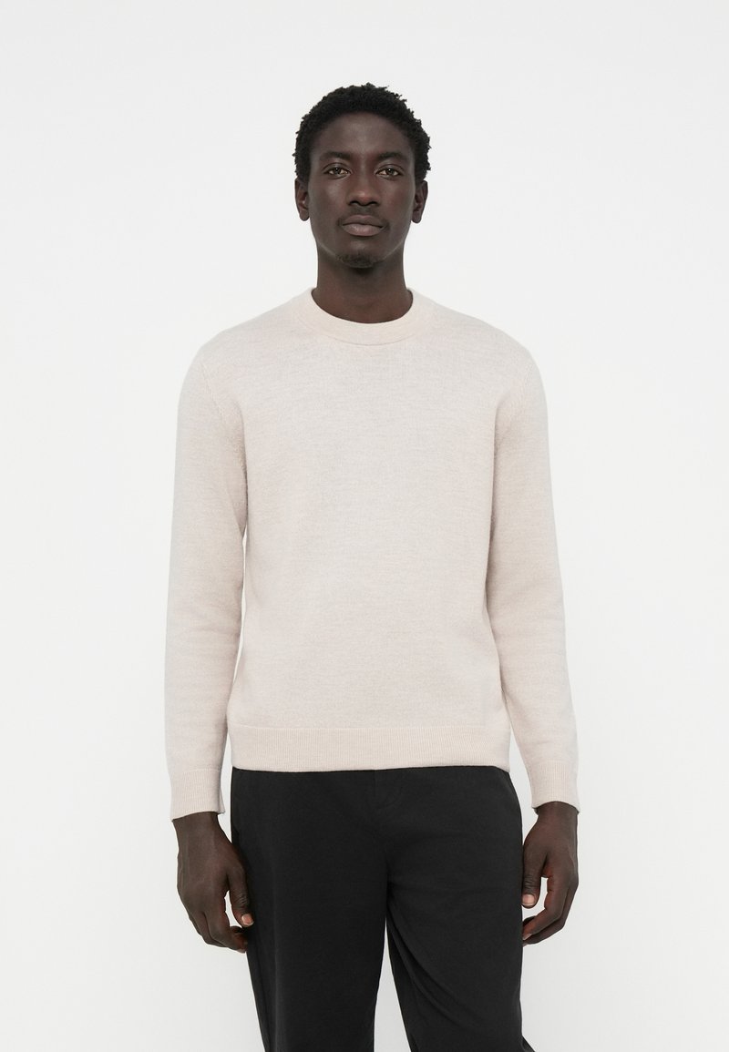 Suéter de punto beige claro con un cuello redondo y mangas largas, que presenta una textura suave y puños y dobladillo acanalados. Combinado con pantalones negros.