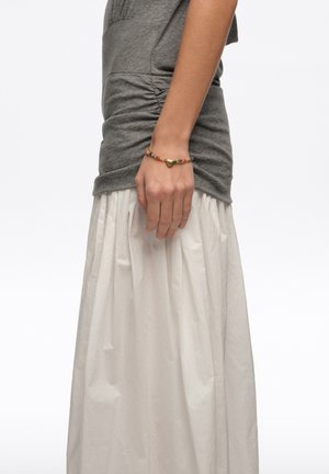 Kobieta nosząca złotą bransoletkę z zawieszką w kształcie serca i wielokolorowymi koralikami, szary marszczony top i białą plisowaną spódnicę, stojąca na tle jednolitego tła.