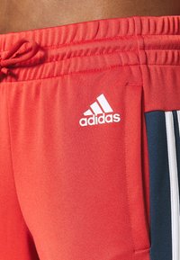 Pantalon de sport rouge Adidas avec taille élastique, cordon de serrage, poche latérale et bandes blanches emblématiques sur un panneau bleu marine.