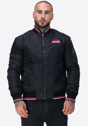 Veste bomber noire en nylon. Caractéristiques : col, poignets et ourlet côtelés avec des rayures rouges et blanches. Poche avec fermeture éclair sur la manche gauche.