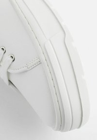 Adidași albi din material sintetic neted, cu vârf rotunjit, talpă groasă și ochiuri metalice. Prezintă cusături minime și șireturi moi.